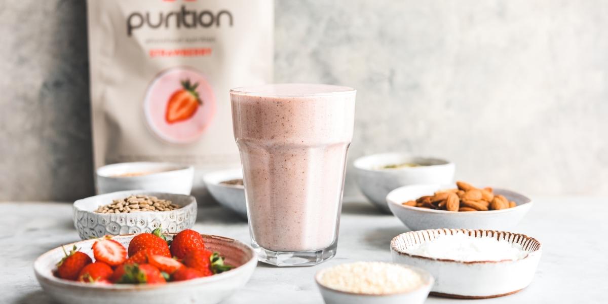 Purition Strwberry meal shake made up in a tall glass surrounded by the whole food ingredients used to make Purition