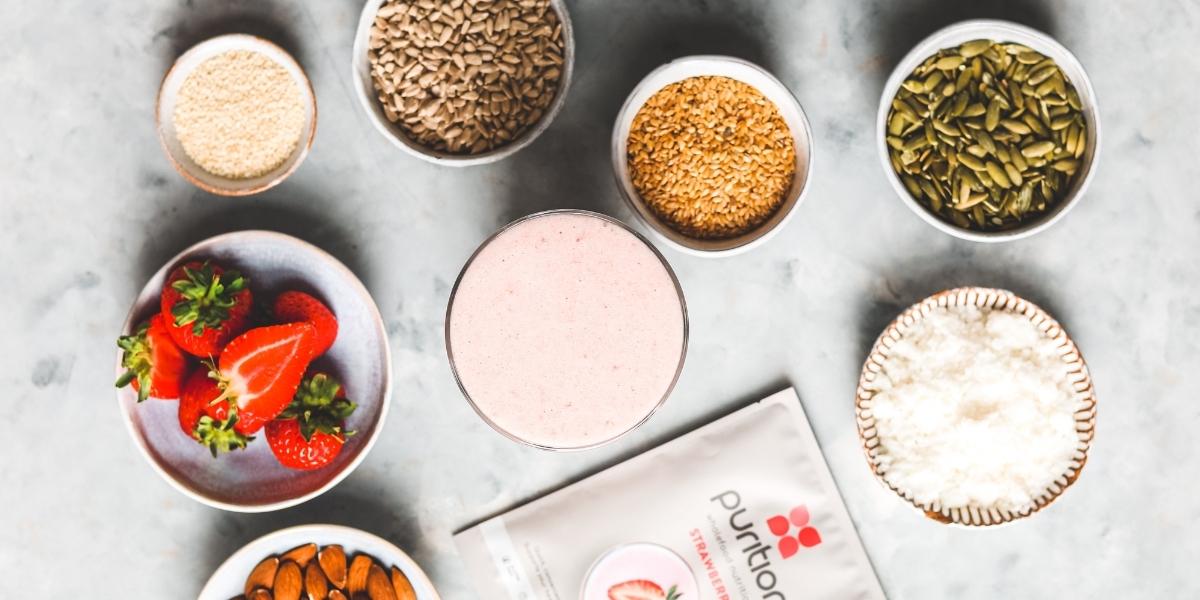 Glass of Purition Vegan Strawberry protein powder surrounded by bowls of fresh ingredients