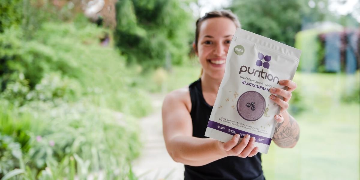 Smiley person holding out a 500g bag of Purition Blackcurrant wholefood nutrition.