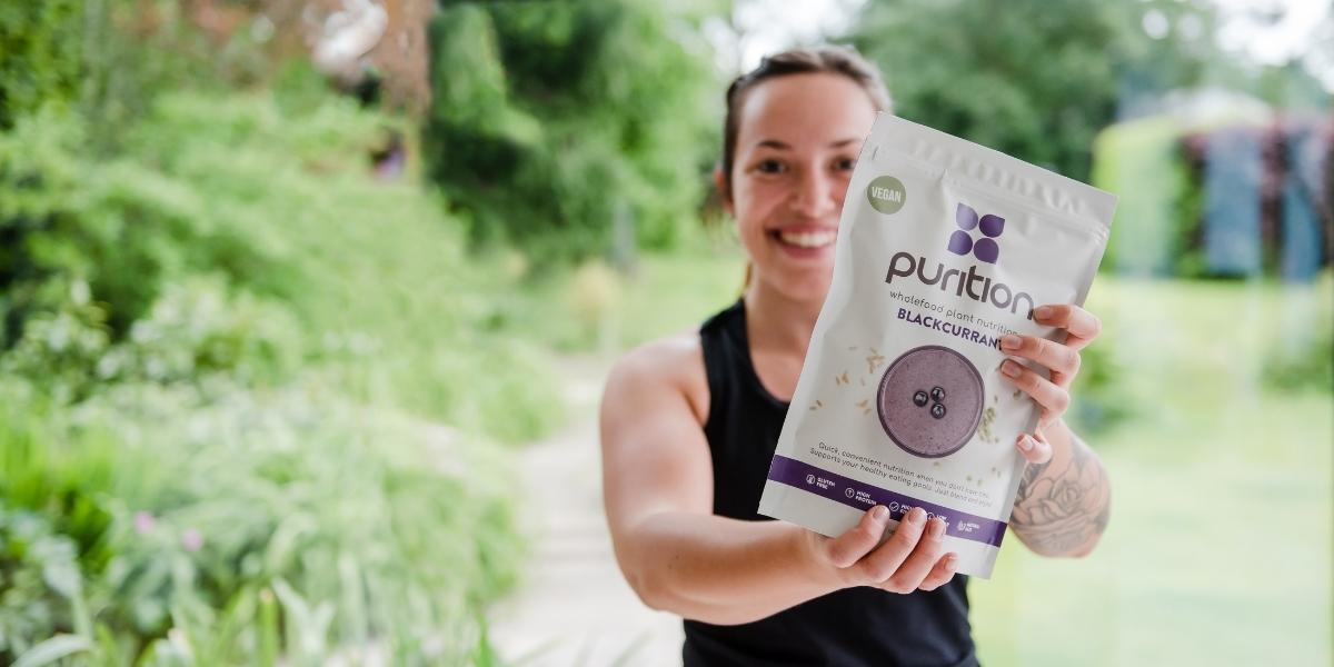 Woman holding Purition Vegan Blackcurrant 500g bag surrounded by greenery