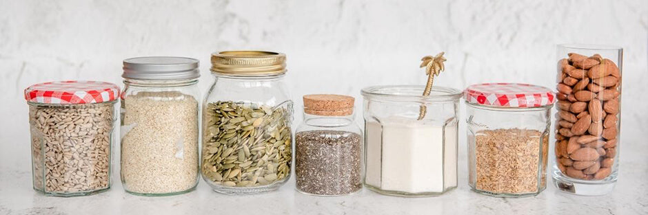 A row of 7 jars containing all the seeds and nuts used to make Purition; sesame seeds, flaxseed, coconut, pumpkin seeds, chia seeds, almonds & sunflower kernels.