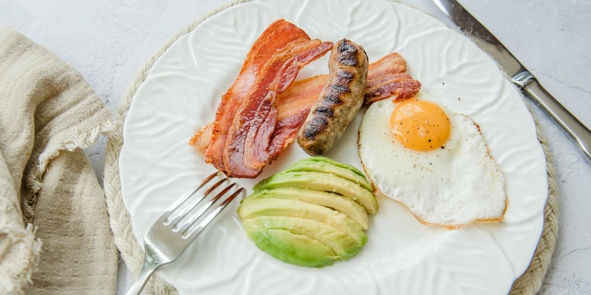 Colourful cooked english breakfast on a plate; grilled crispy bacon and sausage, half a sliced avocado and a fried egg - sunny side up!