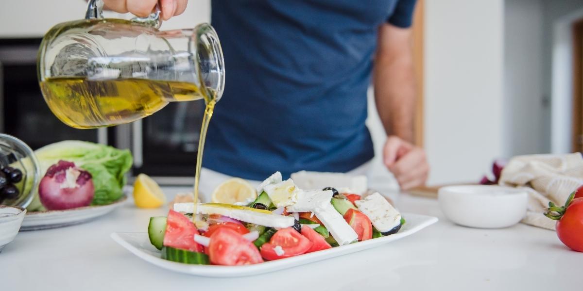 Person dressing a Greek salad with a drizzle of olive oil for a healthy lunch idea.