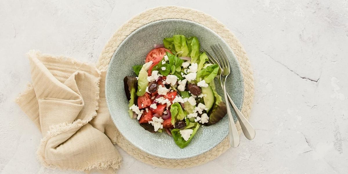 A bowl of colourful Greek salad; greens, tomatoes, olives, red onion and crumbled Feta cheese.