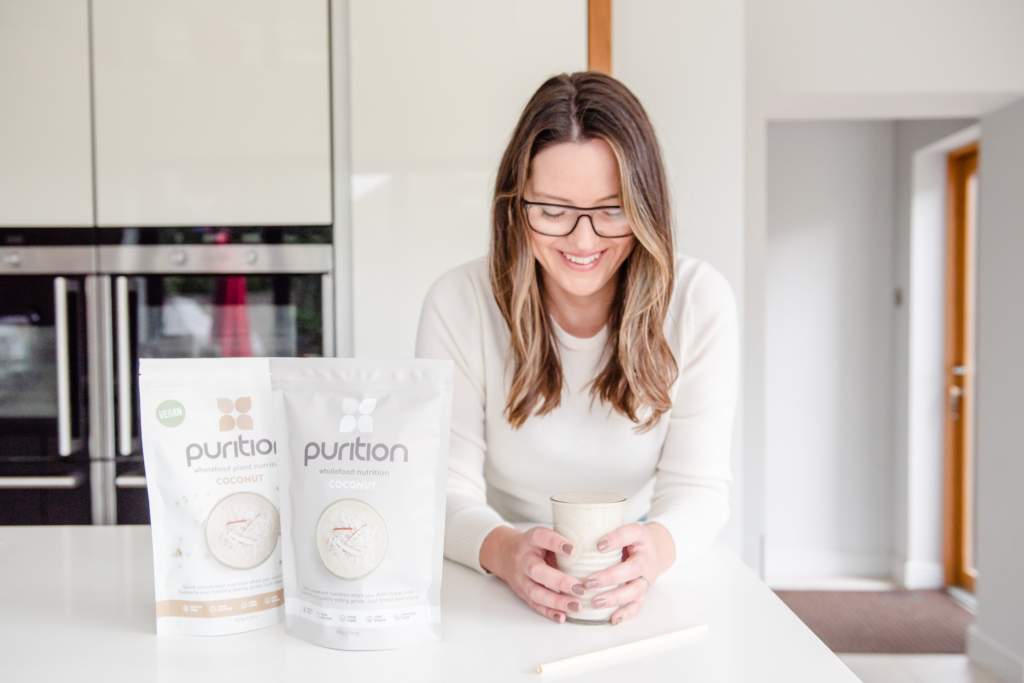 Smiley person in kitchen holding a glass of Purition Coconut wholefood meal shake and 2 bags of Purition Coconut 